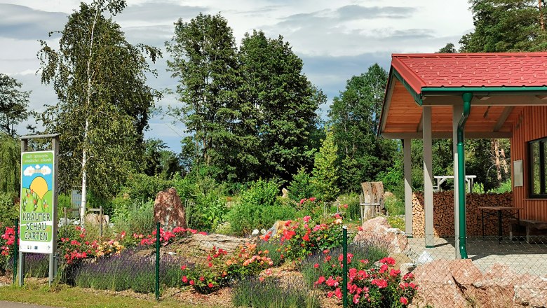 Kräuterschaugarten, © Gemeinde Weinburg Ein Kräuterschaugarten mit bunten Blumen, Bäumen und einem Gebäude mit rotem Dach.