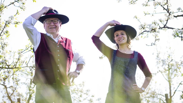 Distelberger, © Martina Schuller Zwei Personen in traditioneller Kleidung mit Hüten stehen in einem blühenden Obstgarten.