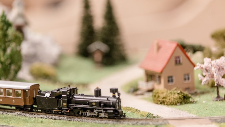 Modellbahn mit Dampflok und Waggon auf einer Miniaturlandschaft mit Haus und Bäumen.
