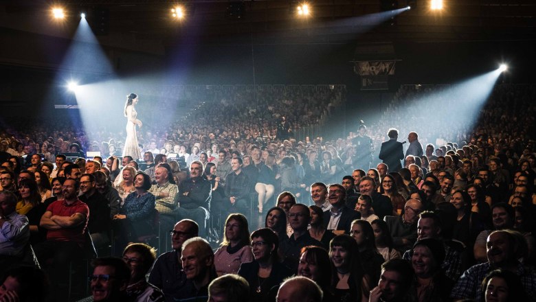 Eine Frau in einem weißen Kleid geht durch ein Publikum in einem Theater oder Konzertsaal, während Scheinwerfer sie beleuchten.
