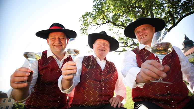 Drei Männer in traditioneller Kleidung mit Hüten halten Weingläser in die Kamera.