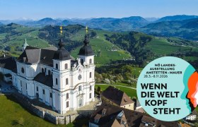Luftaufnahme der Basilika Sonntagberg mit umliegender Landschaft und einem Werbeplakat für die NÖ Landesausstellung 2026.