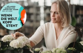 Frau betrachtet Pflanzen in einem Gewächshaus mit einem runden Logo der NÖ Landesausstellung 2026 im Vordergrund.