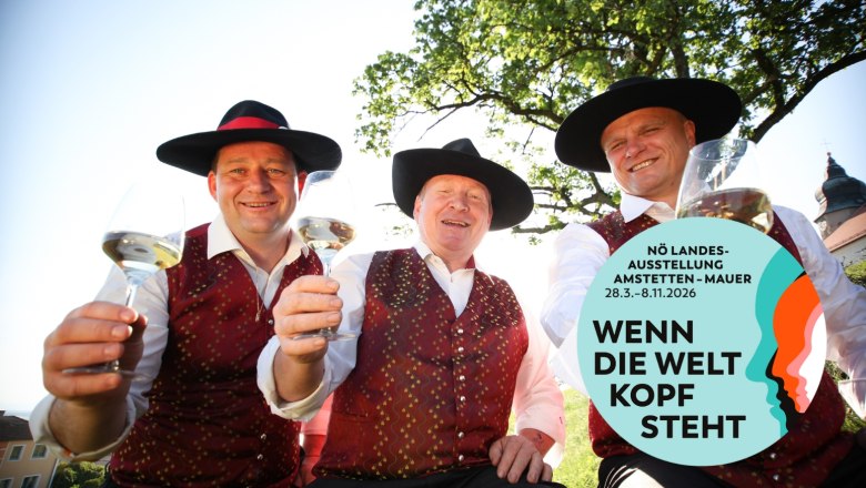 Drei Männer in traditioneller Kleidung mit Weingläsern, lächelnd unter einem Baum. Logo der NÖ Landesausstellung 2026 rechts unten.