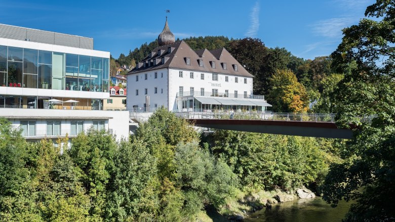 Ein modernes Geb&auml;ude neben einem historischen Schloss, umgeben von B&auml;umen und einem Fluss, unter blauem Himmel.