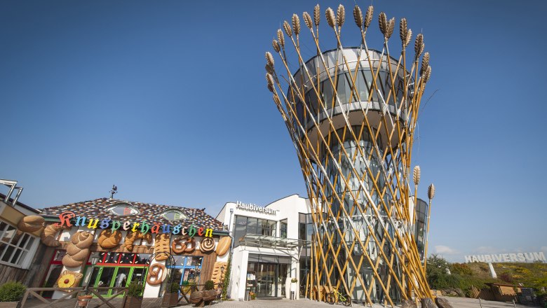 Ein modernes Gebäude mit einem Turm, der von Weizenähren umgeben ist, neben einem dekorativen Knusperhäuschen.