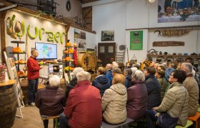 Ein Mann hält einen Vortrag vor einer Gruppe von Menschen in einem rustikalen Raum mit Kürbissen und landwirtschaftlichen Werkzeugen.