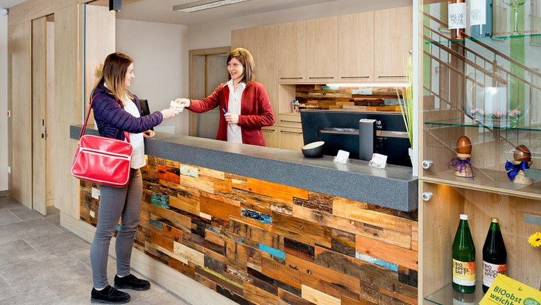 Zwei Frauen am Check-In-Schalter, eine &uuml;berreicht der anderen ein Dokument. Der Schalter ist aus Holz, daneben ein Regal mit Getr&auml;nken und Dekoration.
