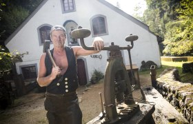 Ein Mann in traditioneller Kleidung steht vor einem historischen Gebäude mit Werkzeugen.