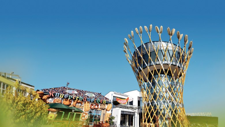 Ein modernes Geb&auml;ude mit einem Turm, der von Weizen&auml;hren umgeben ist, neben einem dekorativen Knusperh&auml;uschen.