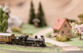 Modellbahn mit Dampflok und Waggon auf einer Miniaturlandschaft mit Haus und Bäumen.